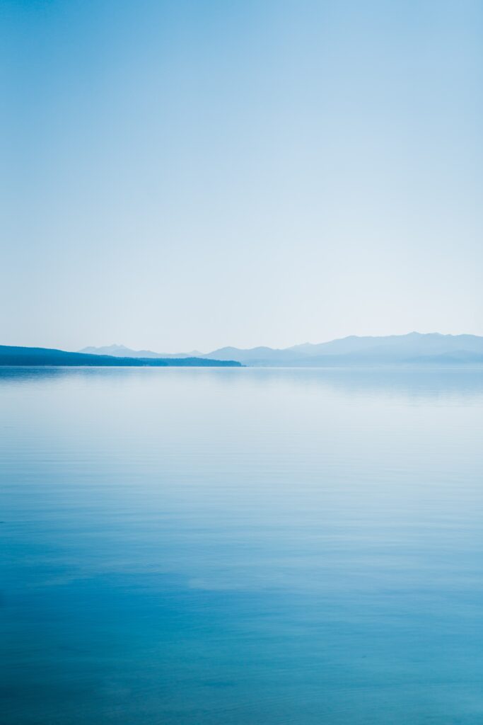Tranquil lake wallpaper with soft blue sky distant mountains trees reflections minimalist landscape