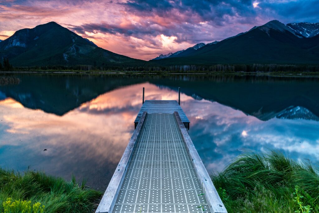Tranquil pier over serene lake with lush grass reflecting dramatic clouds and majestic mountains Wallpaper
