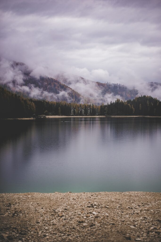 Tranquil lake with sandy shore reflecting misty mountains dense forests and drifting clouds Wallpaper