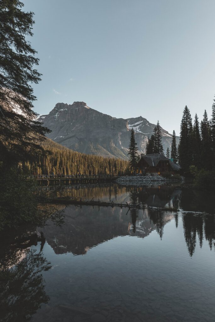 Serene lake reflecting rustic house surrounded by dense forest and towering mountains under clear sky Wallpaper