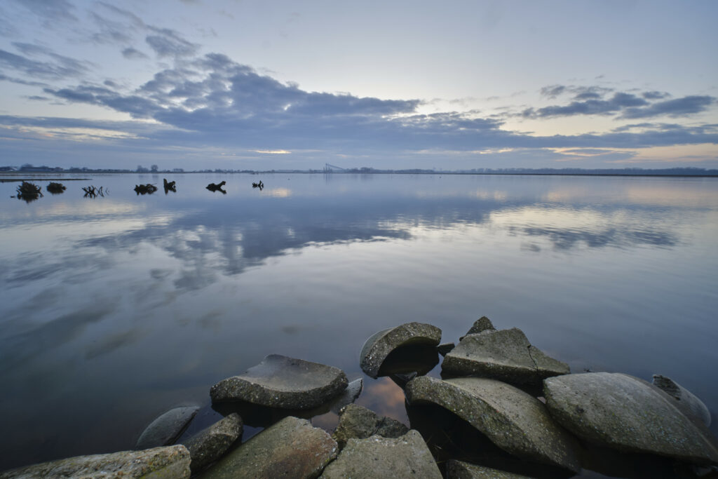 Serene lake reflecting blue gray sky with scattered clouds and concrete fragments foreground wallpaper