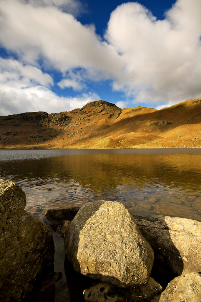 Serene lake with smooth stones shore golden hills and dramatic cloudy sky nature wallpaper background