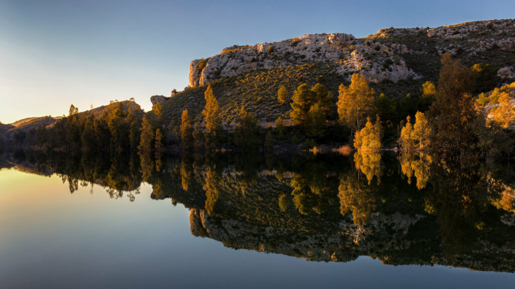 Sunset over tranquil lake reflecting golden trees and rocky hillside serene nature wallpaper