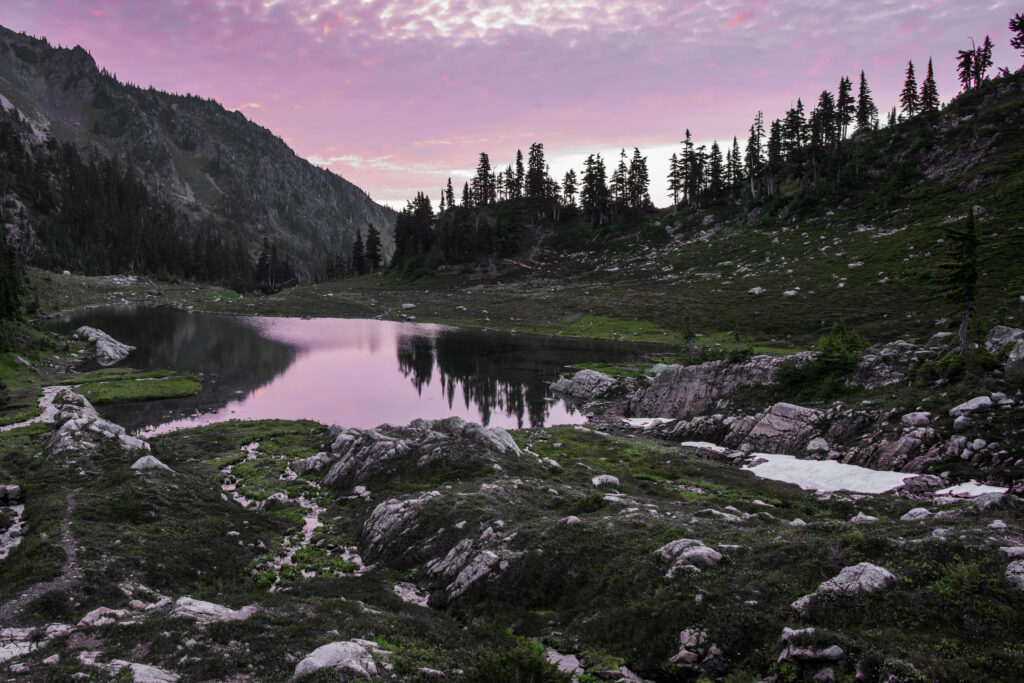 Tranquil lake reflecting pink sunset sky surrounded by rugged mountains and evergreen trees Wallpaper
