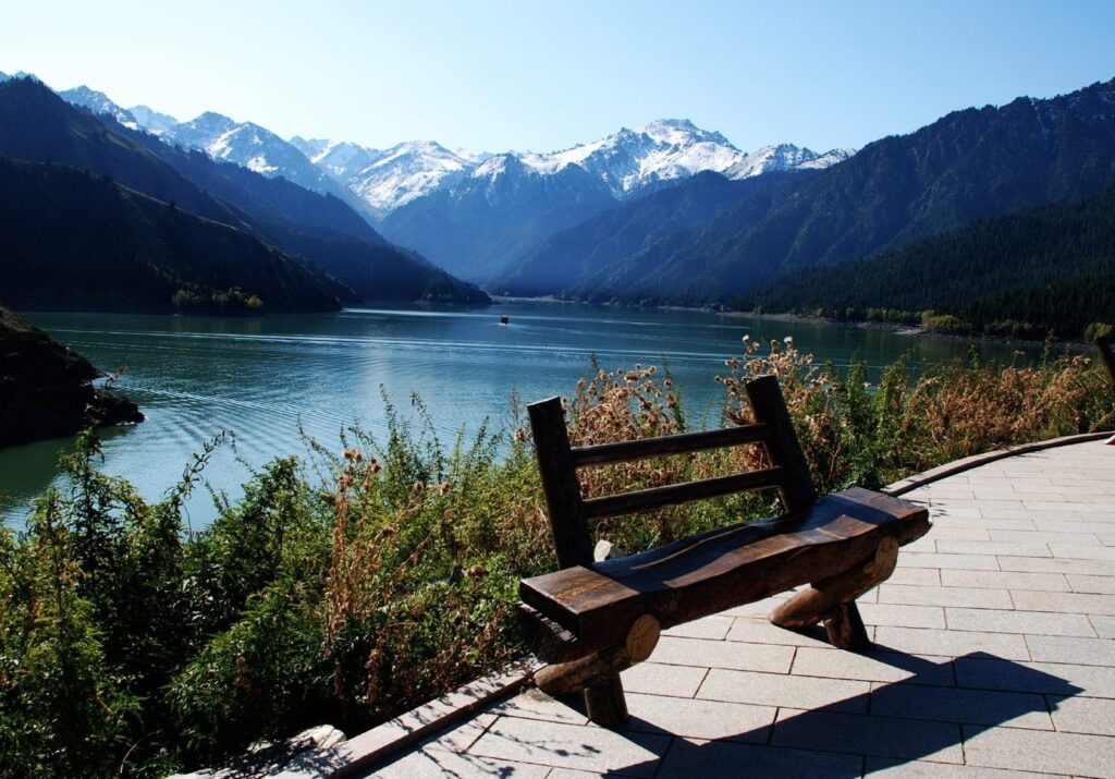 Rustic wooden bench by tranquil lake with lush green plants and snow-capped mountains background Wallpaper