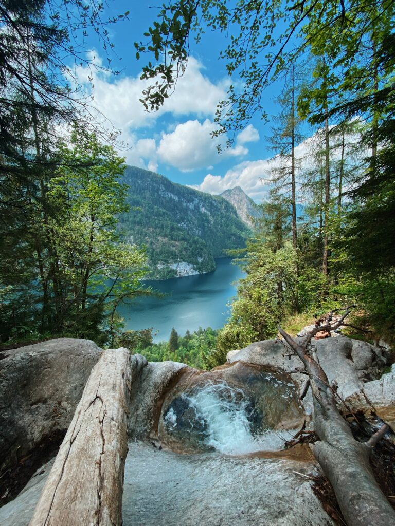 Serene lake with lush trees cliffs and waterfall under vibrant blue sky and scattered clouds wallpaper