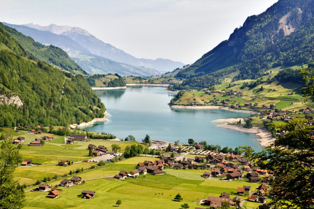 Tranquil lake in green valley with countryside houses and dramatic mountains under clear sky Wallpaper