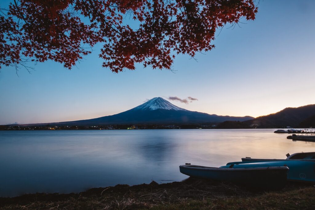 Tranquil lake at dusk reflecting volcano and mountains with autumn leaves and boats on shore Wallpaper