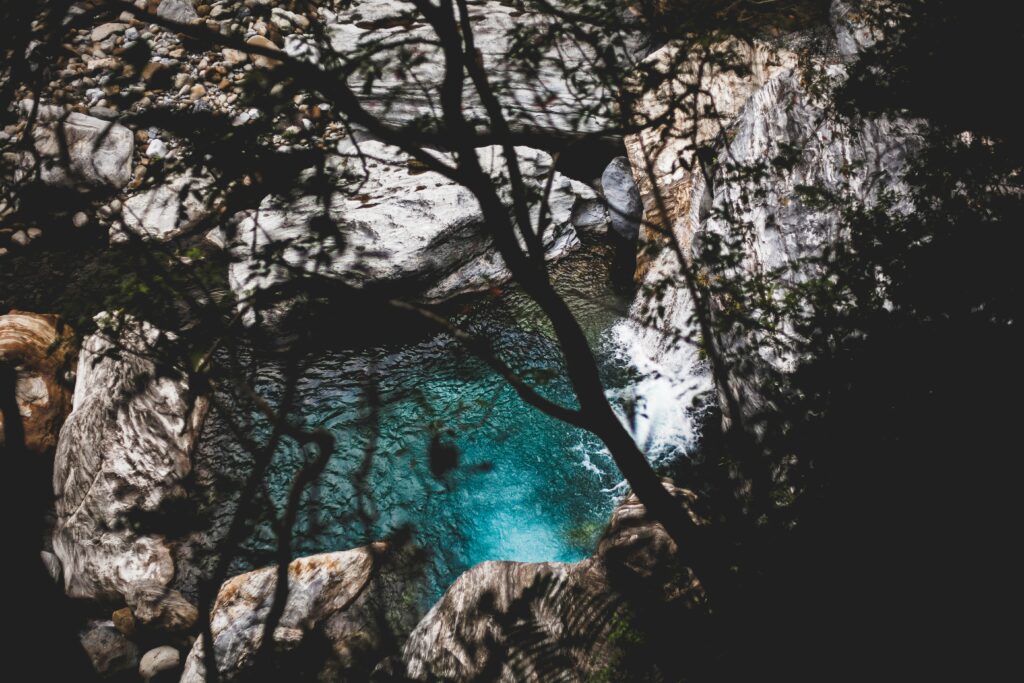 Tranquil lake with turquoise water and rugged stone formations under lush overhanging branches Wallpaper