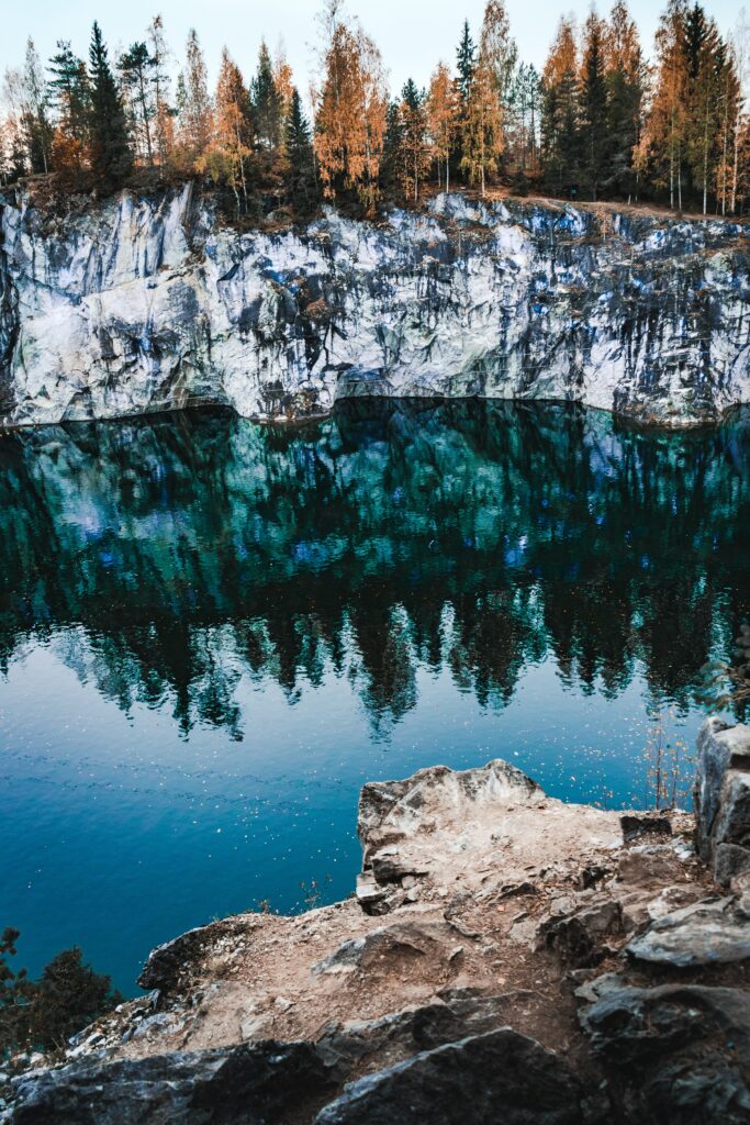Tranquil lakeside with rugged rocks reflecting autumn trees and a towering rocky cliff background Wallpaper