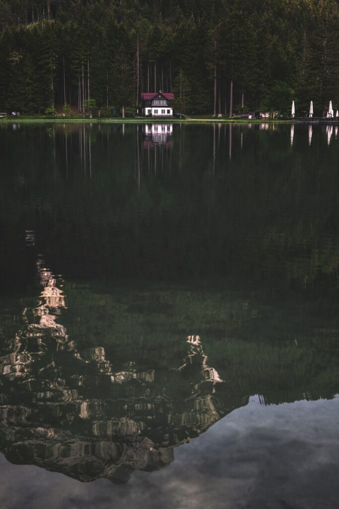 Serene lakeside house surrounded by dense forest with clear water reflecting trees and sky Wallpaper