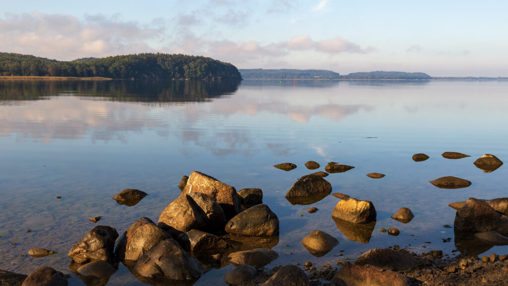 Serene lakeside with smooth stones clear water lush green trees blue sky peaceful nature wallpaper