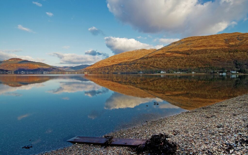 Tranquil reflective lake with pebble shore golden mountains and clear sky serene wallpaper background