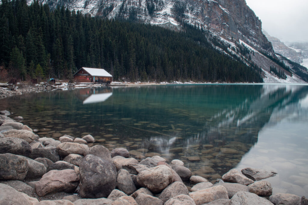 Rustic wooden house by clear lake with smooth stones and misty mountain forest background wallpaper