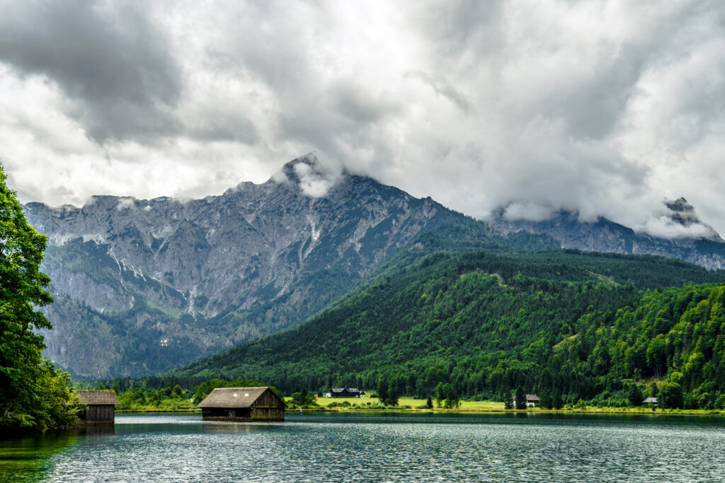 Rustic lakeside house with calm water forest and mountains under dramatic cloudy sky nature wallpaper