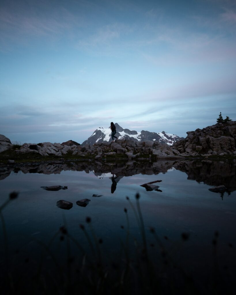 Lone silhouette on rocky lakeshore with snow-capped mountains reflecting in calm evening water Wallpaper
