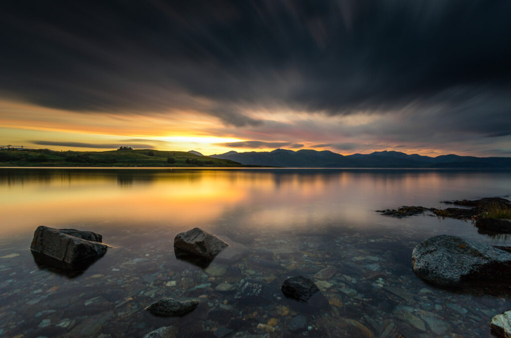 Tranquil lakeside sunrise with smooth stones clear water golden reflections hills and dramatic sky Wallpaper