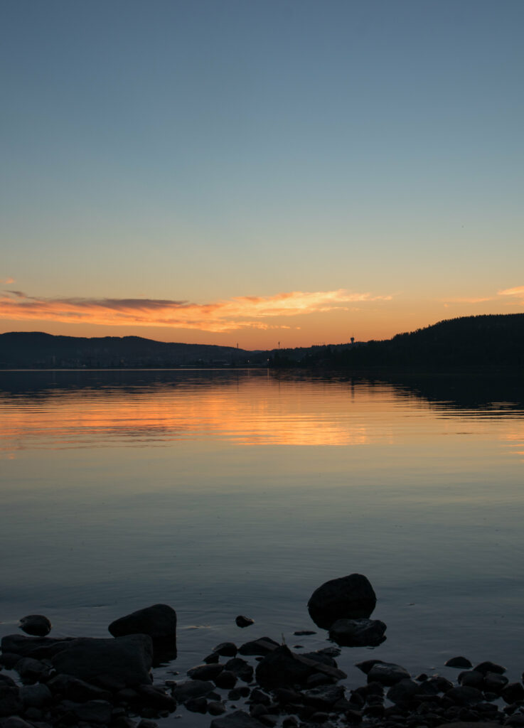 Tranquil lakeside sunset with calm water reflecting orange blue sky and smooth stones foreground Wallpaper