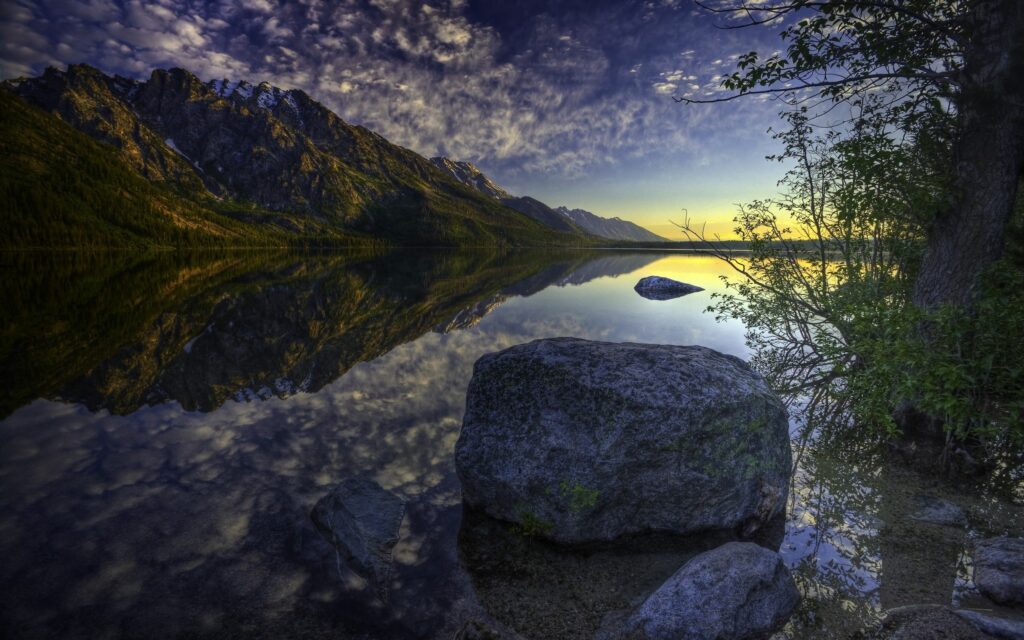 Tranquil lakeside with large gray stone vibrant sunset mountains reflection and lush greenery Wallpaper