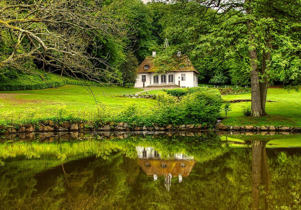 Liselund Slot white manor with thatched roof reflected in calm pond surrounded by lush greenery Wallpaper