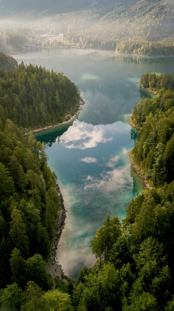 Aerial view of tranquil lake with misty fog surrounded by dense green forest reflecting sky clouds Wallpaper