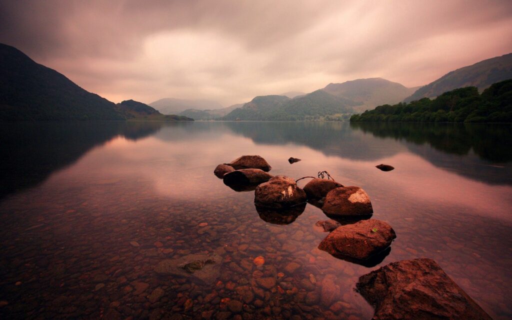 Serene lake with misty mountains smooth rocks under soft overcast sky tranquil nature wallpaper