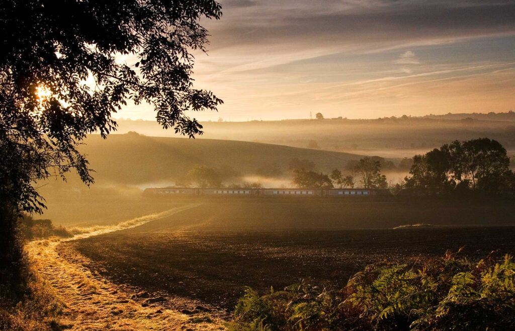Misty morning countryside with winding road train and silhouetted trees under golden sunlight Wallpaper