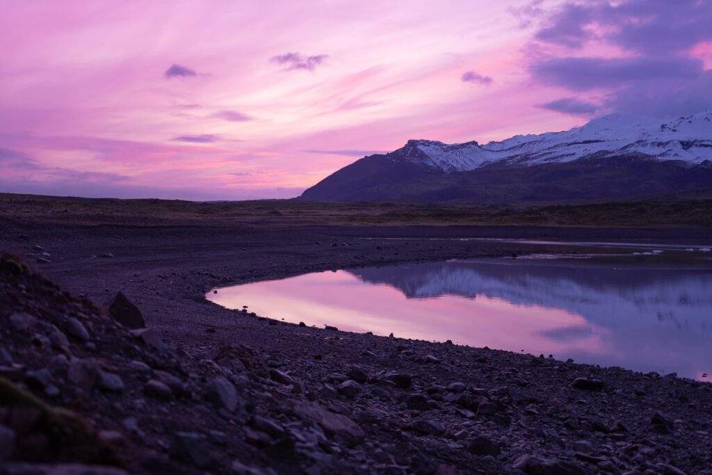 Serene mountain range and calm lake at sunset with purple pink sky and shoreline stones wallpaper