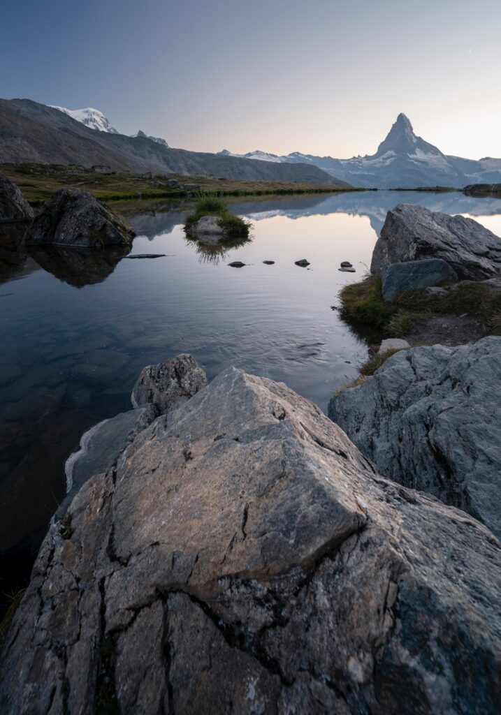 Tranquil lake with rugged stones and majestic mountains under clear soft sky natural landscape wallpaper