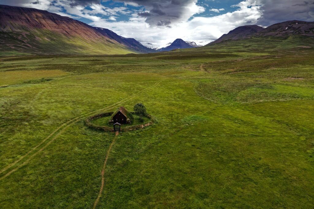 Small hut in green field with scattered rocks and dramatic mountains under cloudy sky wallpaper