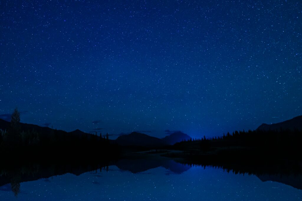 Tranquil mountain landscape with starry sky and tree silhouettes reflected in calm lake at night Wallpaper