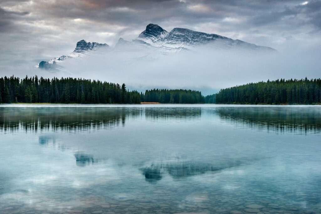 Serene mountain peak reflected in calm lake with misty forest and dramatic cloudy sky background Wallpaper