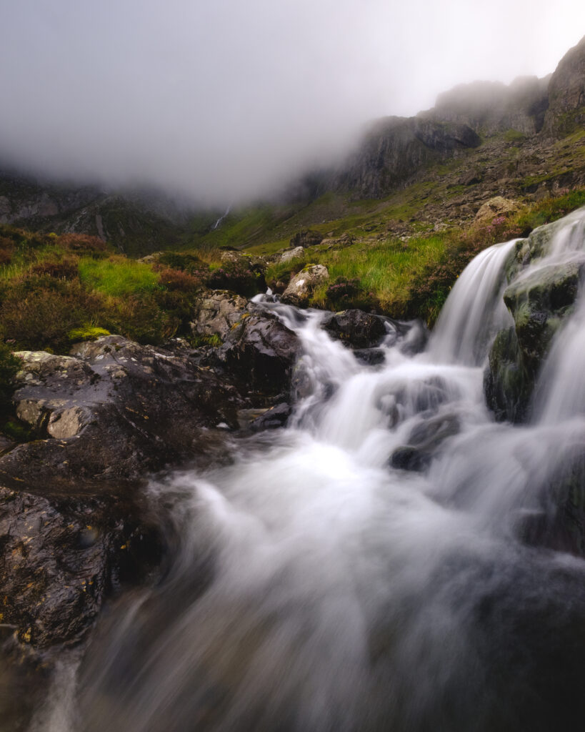 Serene mountain stream flowing over smooth stones with lush grass and misty peaks background Wallpaper