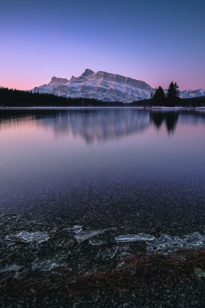 Photorealistic mountain lake at sunset with purple pink sky rocky shore and silhouetted trees Wallpaper