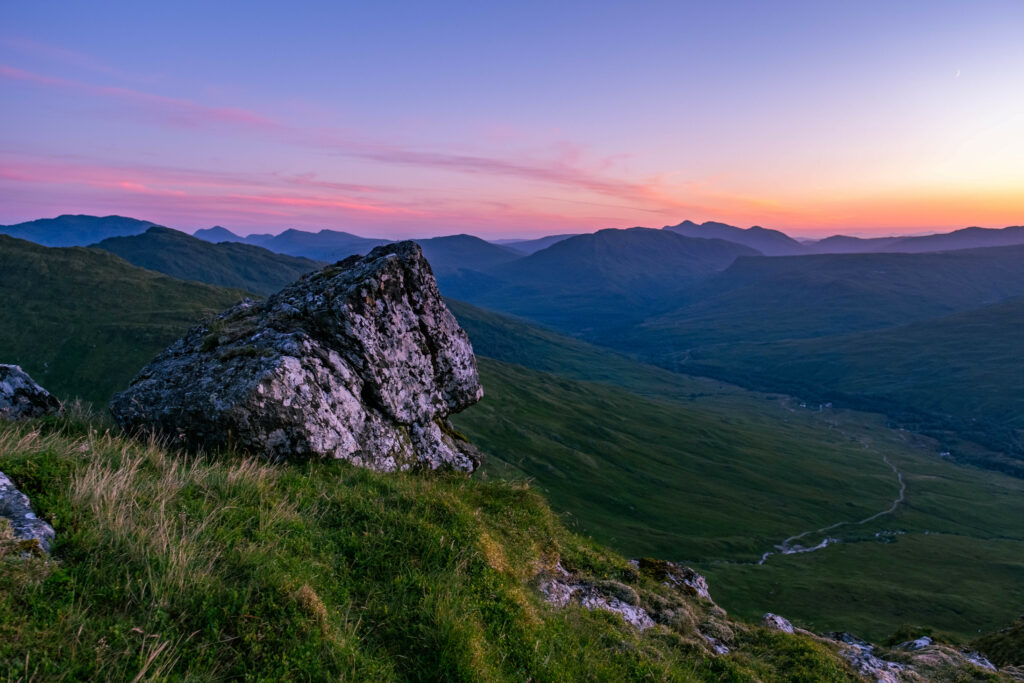 Rugged stone on grassy slope overlooking green valley and layered mountains under evening sky Wallpaper
