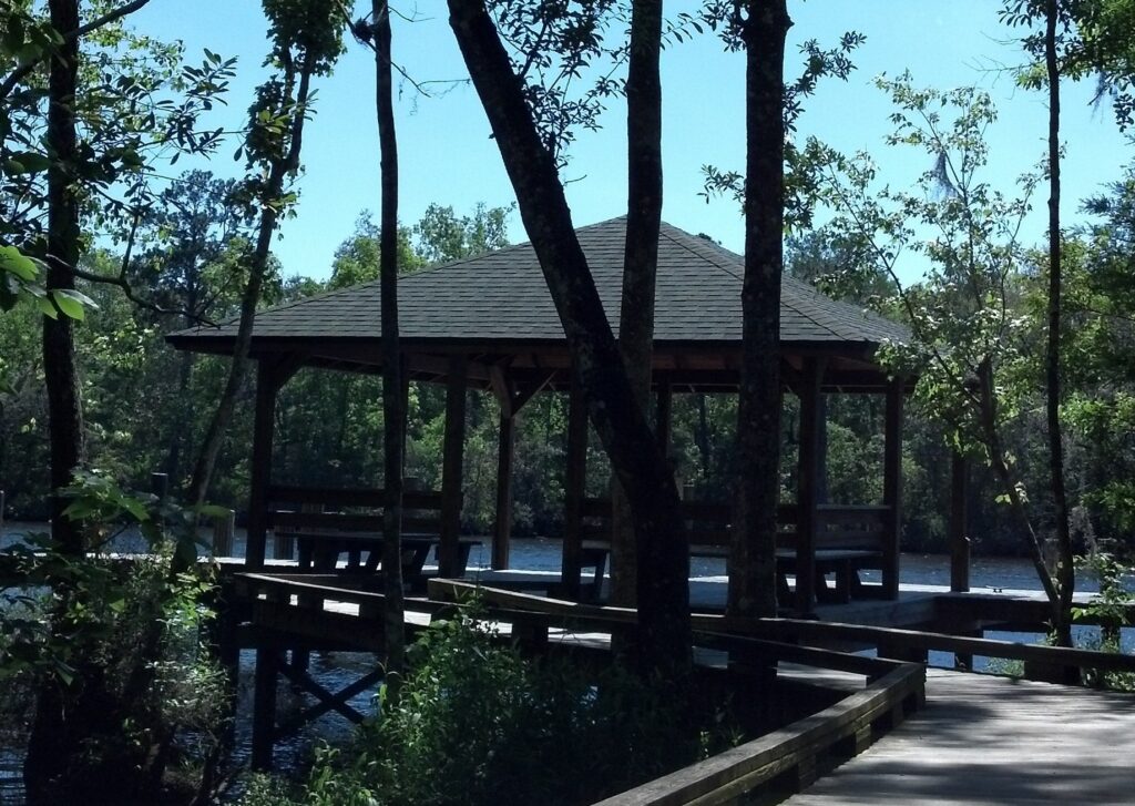 Tranquil dock with wooden pathway and pavilion surrounded by lush green trees and calm water Wallpaper