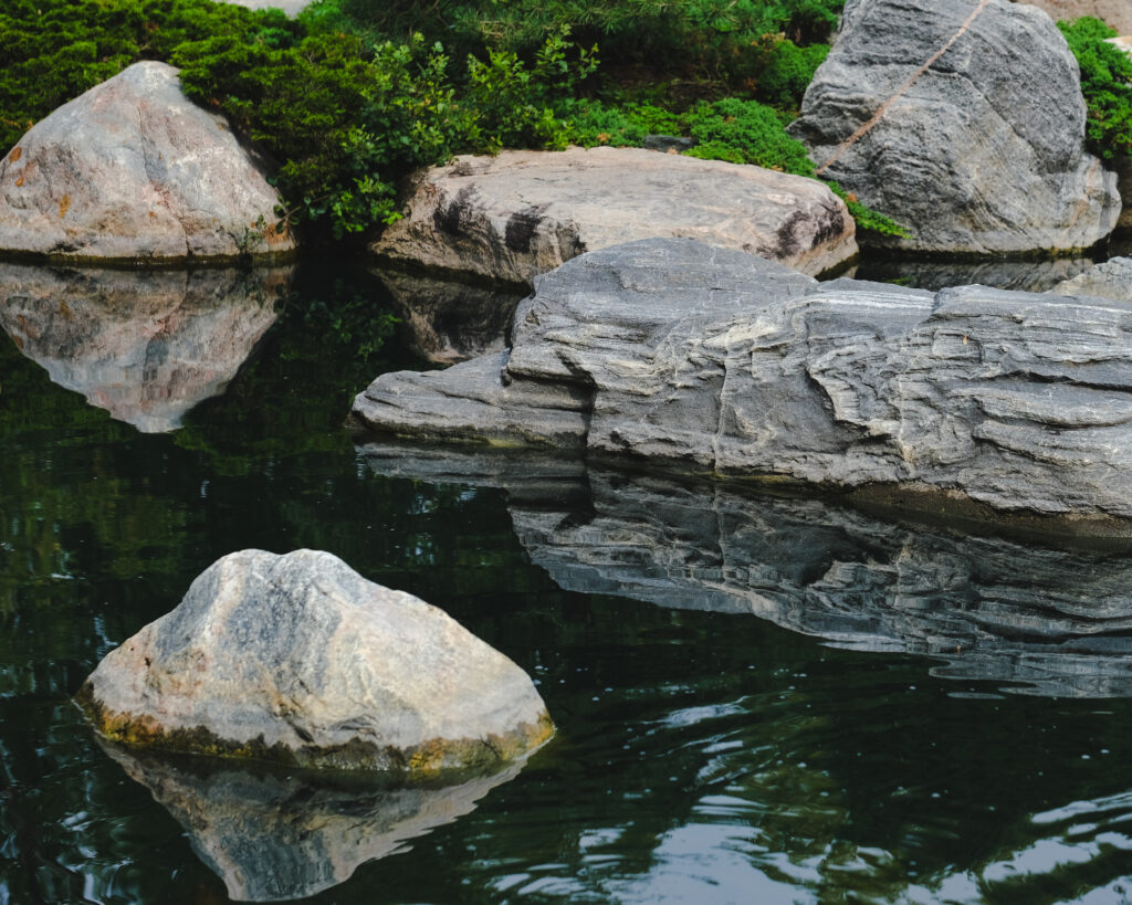 Large stones by calm pond with clear reflections surrounded by green foliage peaceful nature wallpaper