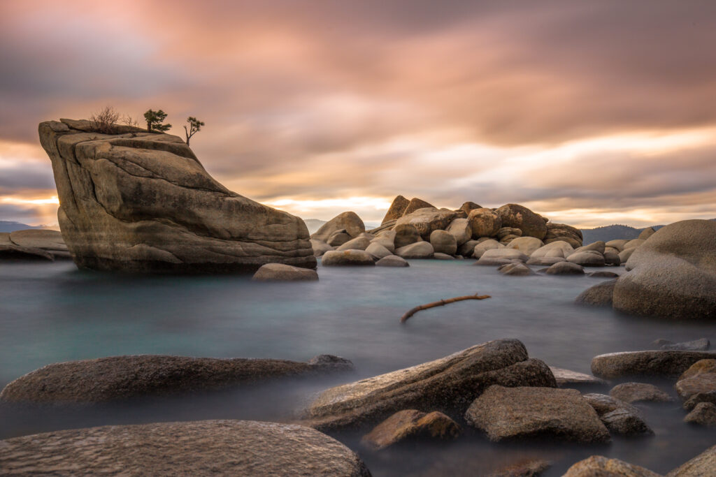Calm turquoise water with smooth stones and a floating branch under colorful cloudy sky wallpaper