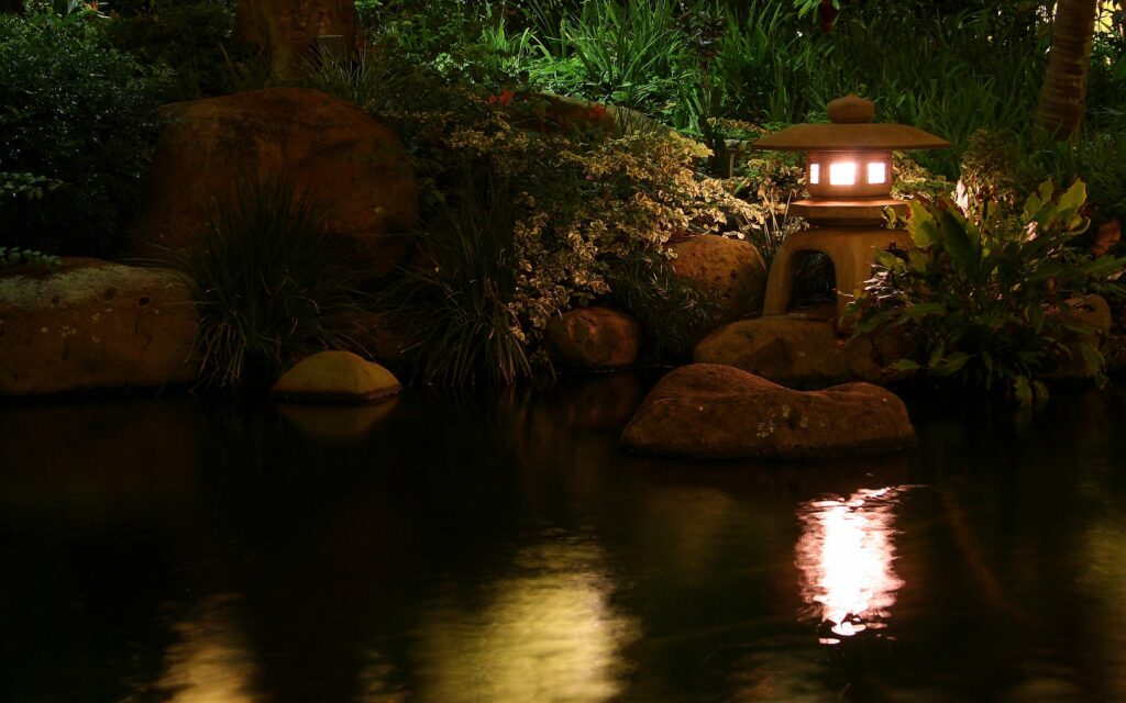 Traditional Chinese stone lamp glowing by pond with reflections lush plants smooth stones serene night Wallpaper