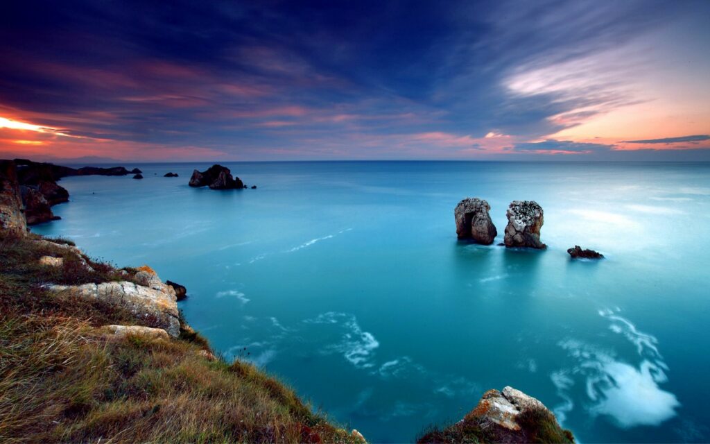Sunset over tranquil ocean gulf with smooth blue water lilac sky and rugged coastal rocks Wallpaper