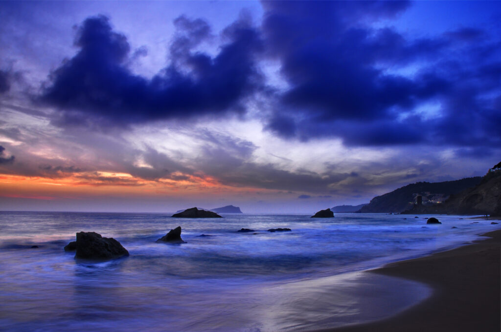 Tranquil ocean sunset with scattered shore stones vibrant blues purples and warm orange hues Wallpaper