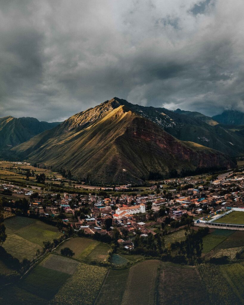 Aerial view of serene Peruvian village with green fields and sunlit mountains under moody sky Wallpaper