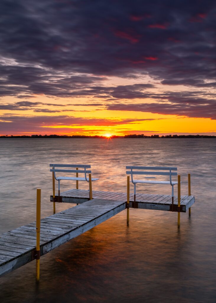 Tranquil pier with white benches over calm sea at vibrant sunset with orange purple sky clouds Wallpaper