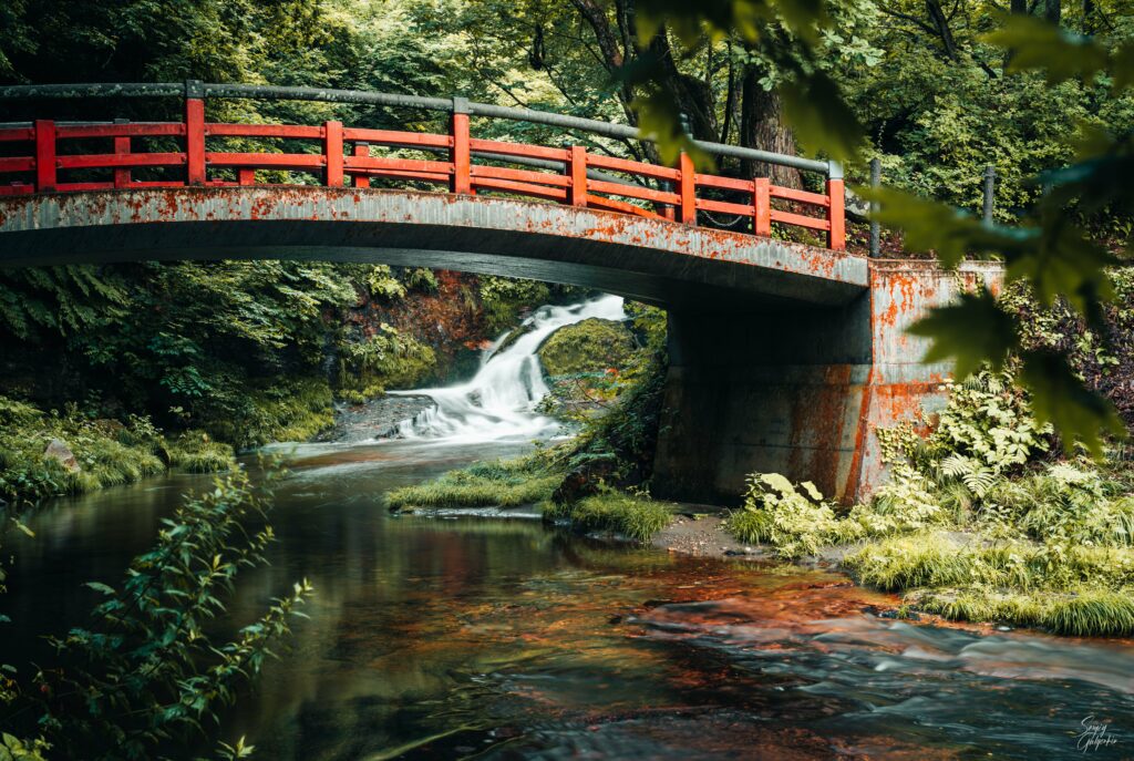 Vibrant red bridge over tranquil river with lush greenery and gentle waterfall serene nature scene Wallpaper