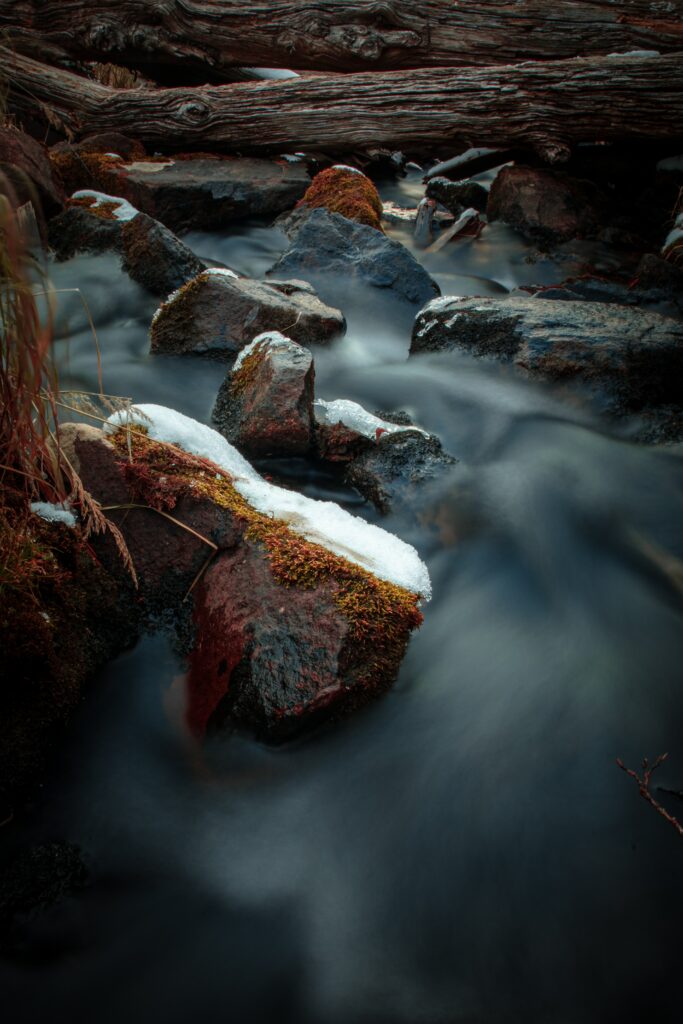 Serene river flowing around mossy stones and logs with snow and soft natural light wallpaper