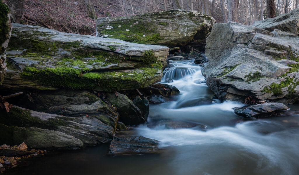 Tranquil river flowing gently through mossy rocks in serene natural landscape with earthy tones Wallpaper
