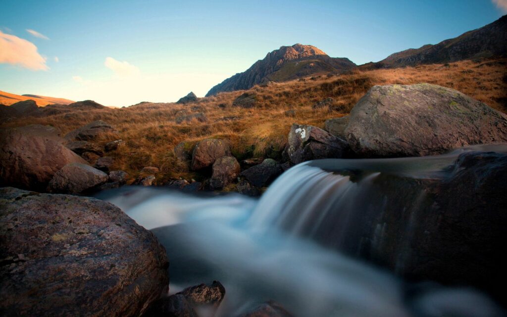 Serene river flowing over smooth rocks with rugged boulders golden grass and distant mountains Wallpaper
