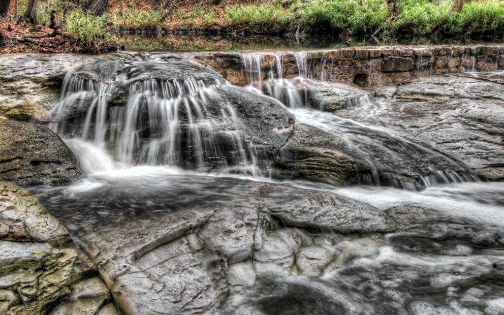 Smooth stones and flowing river water with lush green plants in a tranquil natural wallpaper background