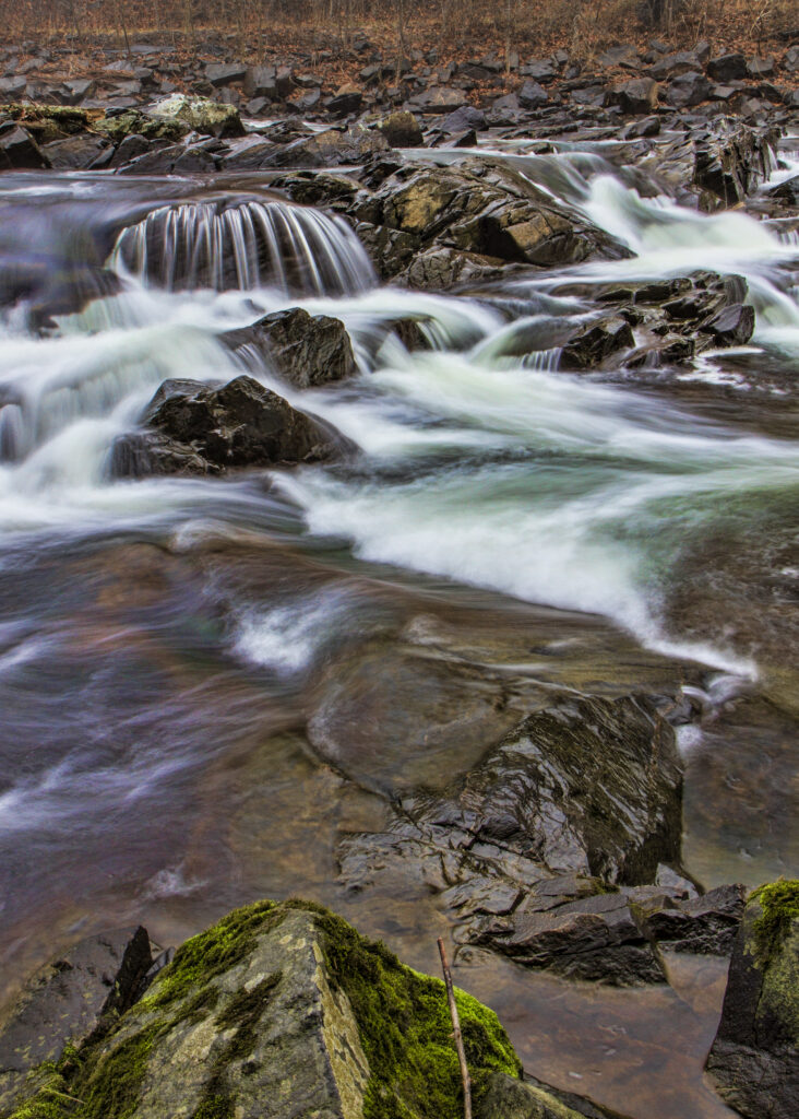 Tranquil river flowing over smooth stones with moss-covered rocks in a serene natural setting Wallpaper