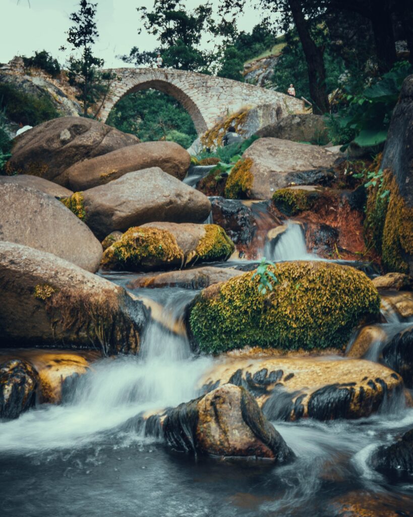 Tranquil river flowing over mossy stones beneath historic stone bridge in lush green forest wallpaper